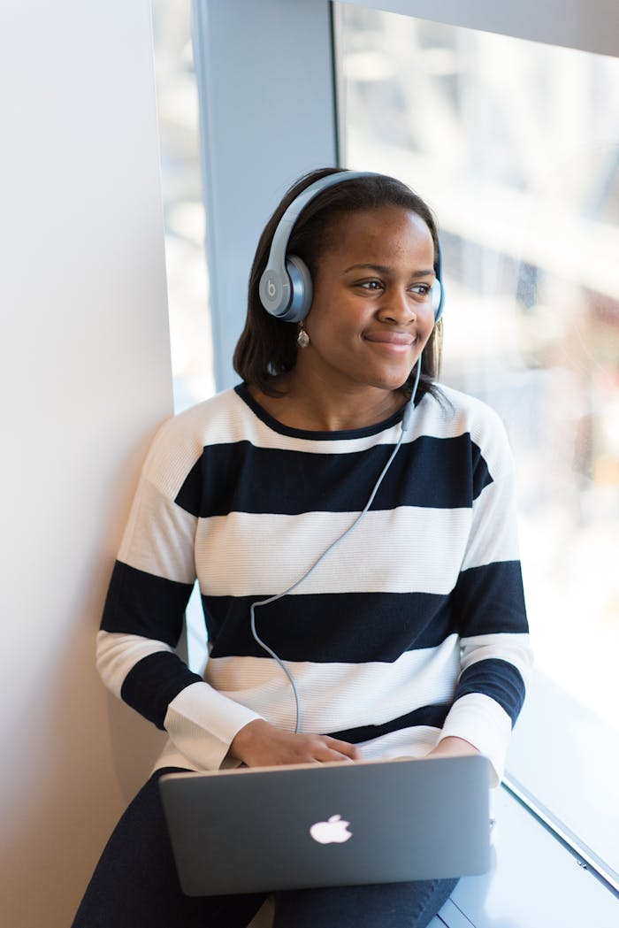 Smiling African American woman listening to music on headphones while using a laptop by a window.